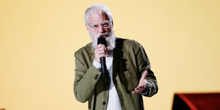 David Letterman in an olive green jacket speaks with his hands on stage