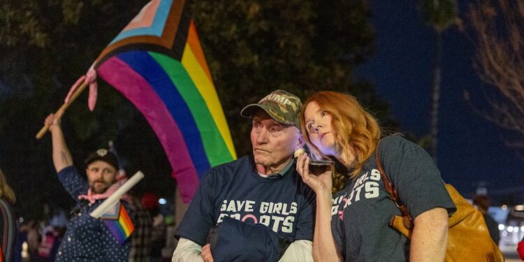 Transgender athlete supporter Kyle Harp, left, of Riverside holds the progress  pride flag as "Save Girls Sports" supporters Lori Lopez and her dad Pete Pickering, both of Riverside, listen to the debate as they join the overflow crowd converging outside the Riverside Unified School District meeting Thursday night to debate the rights of transgender athletes to compete in high school sports Thursday, Dec. 19, 2024. 