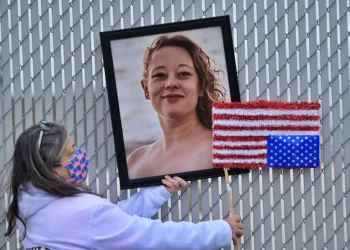 A woman in a mask holds up a portrait of Renee Nicole Good and an upside down American flag in front of a chain link fence.