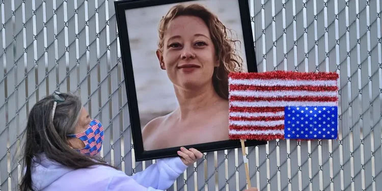 A woman in a mask holds up a portrait of Renee Nicole Good and an upside down American flag in front of a chain link fence.