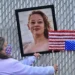 A woman in a mask holds up a portrait of Renee Nicole Good and an upside down American flag in front of a chain link fence.