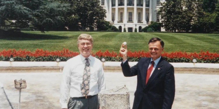 Assistant Curator William G. Allman and Gary J. Walters hold a gold ball and laugh in front of the White House.