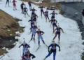 Several biathletes ski on a narrow strip of snow that has grass and mud on either side of it.