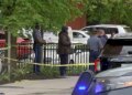 Police officers stand near yellow crime scene tape along a sidewalk in Cambridge, Massachusetts, as vehicles and bystanders are visible nearby.
