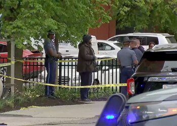 Police officers stand near yellow crime scene tape along a sidewalk in Cambridge, Massachusetts, as vehicles and bystanders are visible nearby.