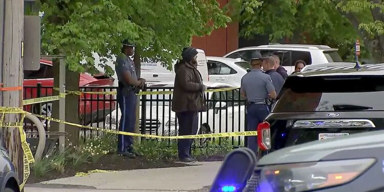 Police officers stand near yellow crime scene tape along a sidewalk in Cambridge, Massachusetts, as vehicles and bystanders are visible nearby.
