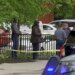 Police officers stand near yellow crime scene tape along a sidewalk in Cambridge, Massachusetts, as vehicles and bystanders are visible nearby.