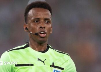 Omar Artan refereeing a match, with earpiece and microphone, and a Fifa badge on his light green shirt