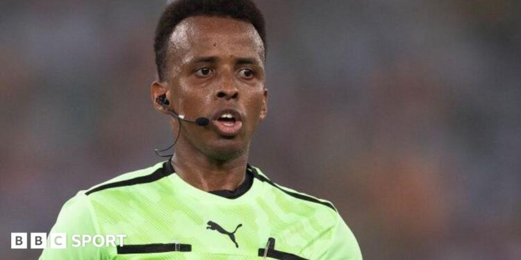 Omar Artan refereeing a match, with earpiece and microphone, and a Fifa badge on his light green shirt