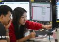 A woman in a red cardigan points to a screen during a discussion with a colleague in a trading room in Seoul