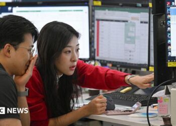 A woman in a red cardigan points to a screen during a discussion with a colleague in a trading room in Seoul
