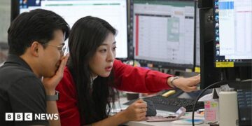 A woman in a red cardigan points to a screen during a discussion with a colleague in a trading room in Seoul