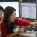 A woman in a red cardigan points to a screen during a discussion with a colleague in a trading room in Seoul