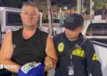 Simon Peter Carman, a man with short light brown hair wearing a sleeveless black too, is escorted by a Thai police officer.