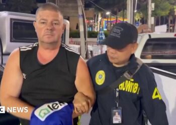 Simon Peter Carman, a man with short light brown hair wearing a sleeveless black too, is escorted by a Thai police officer.
