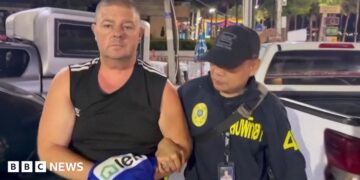 Simon Peter Carman, a man with short light brown hair wearing a sleeveless black too, is escorted by a Thai police officer.