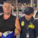 Simon Peter Carman, a man with short light brown hair wearing a sleeveless black too, is escorted by a Thai police officer.