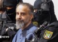 Taleb al-Abdulmohsen, a man with greying brown hair and a greying beard, looks on while being escorted by armed security officers. He wears a light blue buttoned shirt. The security officials wear all-black uniforms, including face coverings with only the eyes visible.