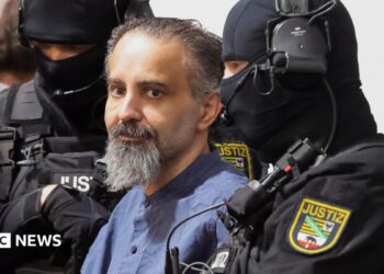 Taleb al-Abdulmohsen, a man with greying brown hair and a greying beard, looks on while being escorted by armed security officers. He wears a light blue buttoned shirt. The security officials wear all-black uniforms, including face coverings with only the eyes visible.