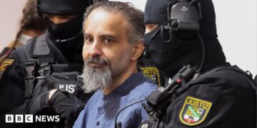 Taleb al-Abdulmohsen, a man with greying brown hair and a greying beard, looks on while being escorted by armed security officers. He wears a light blue buttoned shirt. The security officials wear all-black uniforms, including face coverings with only the eyes visible.