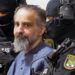 Taleb al-Abdulmohsen, a man with greying brown hair and a greying beard, looks on while being escorted by armed security officers. He wears a light blue buttoned shirt. The security officials wear all-black uniforms, including face coverings with only the eyes visible.