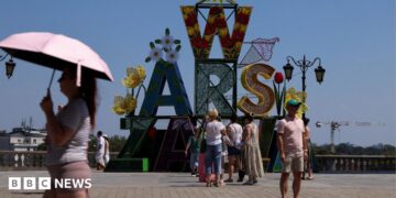 A woman walks under an umbrella past a "Warsaw" sign as a heatwave sweeps Poland, in Warsaw, Poland,