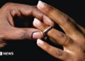 A man places a silver wedding band onto a young woman's ring finger.