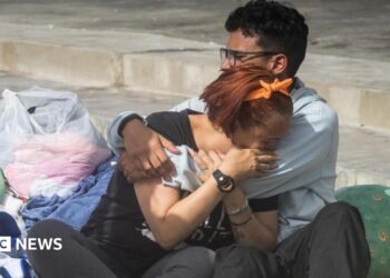 Two people cry into each others' arms surrounded by pillows, blankets and carrier bags of possessions on concrete stairs outside