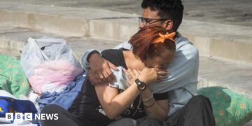 Two people cry into each others' arms surrounded by pillows, blankets and carrier bags of possessions on concrete stairs outside