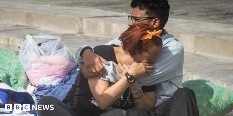 Two people cry into each others' arms surrounded by pillows, blankets and carrier bags of possessions on concrete stairs outside