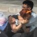 Two people cry into each others' arms surrounded by pillows, blankets and carrier bags of possessions on concrete stairs outside