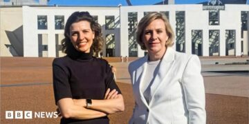 Two women standing outside federal parliament house in Canberra. One is a brunette wearing a black top and with her hands folded. The other has a short blonde bob and wearing a white blazer.