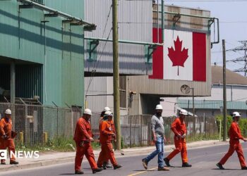 A Canadian national flag is seen in the background as seven workers dressed in orange work suits and white hard hats cross the street in front of ArcelorMittal Dofasco's steel manufacturing buildings in Hamilton, Ontario.