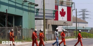 A Canadian national flag is seen in the background as seven workers dressed in orange work suits and white hard hats cross the street in front of ArcelorMittal Dofasco's steel manufacturing buildings in Hamilton, Ontario.
