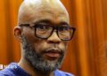 A head and shoulders picture of Cat Matlala in court - he is wearing a blue jumper, the neckline of which can just be seen at the bottom of the picture.