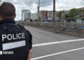 Back of officer in black vest marked with white letters reading "POLICE" next to an empty roadway with buildings across the street