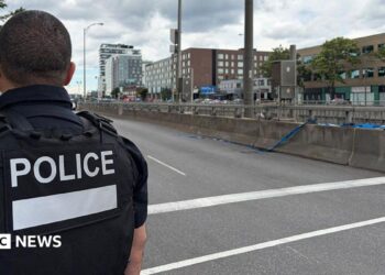 Back of officer in black vest marked with white letters reading "POLICE" next to an empty roadway with buildings across the street
