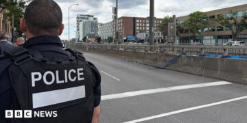 Back of officer in black vest marked with white letters reading "POLICE" next to an empty roadway with buildings across the street