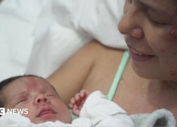 Dayana Patino grins as she holds her son Juan David close to her chest from a hospital bed. Her husband, Gerson, stands beside them wearing a light blue t-shirt.