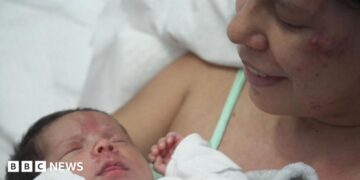 Dayana Patino grins as she holds her son Juan David close to her chest from a hospital bed. Her husband, Gerson, stands beside them wearing a light blue t-shirt.