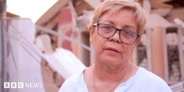 Zuly Marín, dressed in a white shirt and black glasses, stares into space.