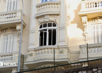 The damaged window of a light yellow residential building, which has balconies and ornate carvings.