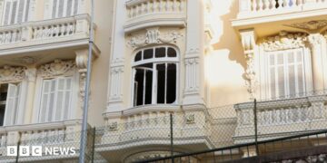 The damaged window of a light yellow residential building, which has balconies and ornate carvings.