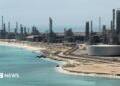 A file photo of Aramco's Ras Tanura oil refinery and oil terminal, which lies along a stretch of coast. The site comprises a network of several tall chimney-shaped structures and smaller buildings. It is daytime and the water along the coastline is a clear blue.