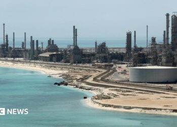 A file photo of Aramco's Ras Tanura oil refinery and oil terminal, which lies along a stretch of coast. The site comprises a network of several tall chimney-shaped structures and smaller buildings. It is daytime and the water along the coastline is a clear blue.