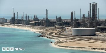 A file photo of Aramco's Ras Tanura oil refinery and oil terminal, which lies along a stretch of coast. The site comprises a network of several tall chimney-shaped structures and smaller buildings. It is daytime and the water along the coastline is a clear blue.