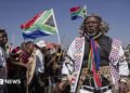 Hundreds of protesters marching outdoors, chanting and holding south african flags and signs