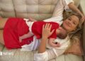 A woman with long blonde hair, wearing a white top and red skirt lying on a cream couch, with a baby sleeping on her chest.