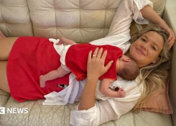 A woman with long blonde hair, wearing a white top and red skirt lying on a cream couch, with a baby sleeping on her chest.