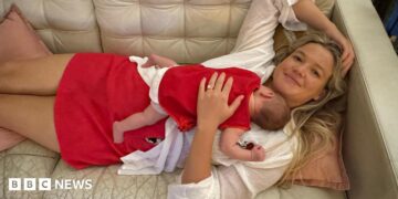 A woman with long blonde hair, wearing a white top and red skirt lying on a cream couch, with a baby sleeping on her chest.